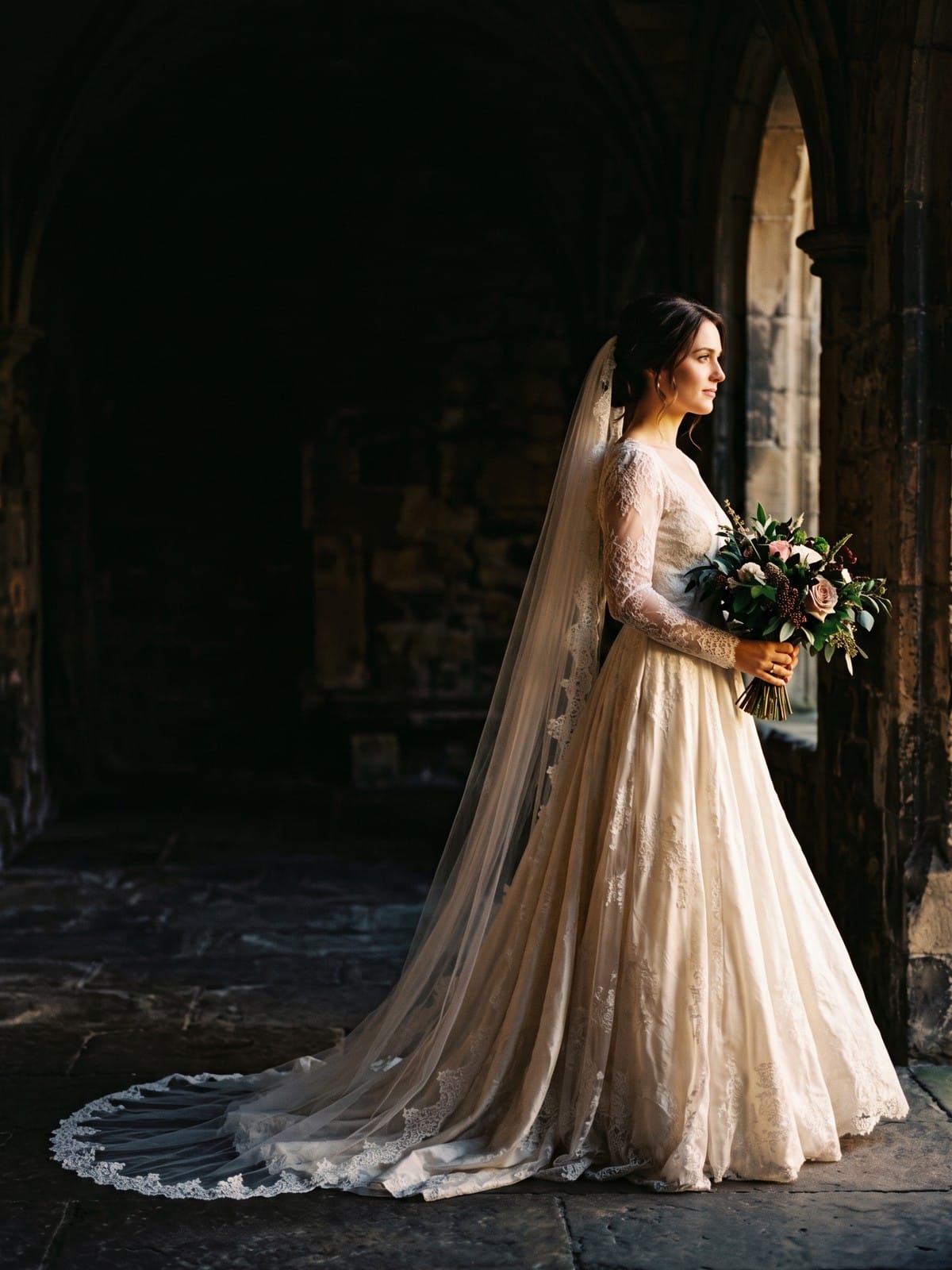 A bride in a lace gown and cathedral veil stands in a stone cloister, holding her bouquet in the light from an arched window
