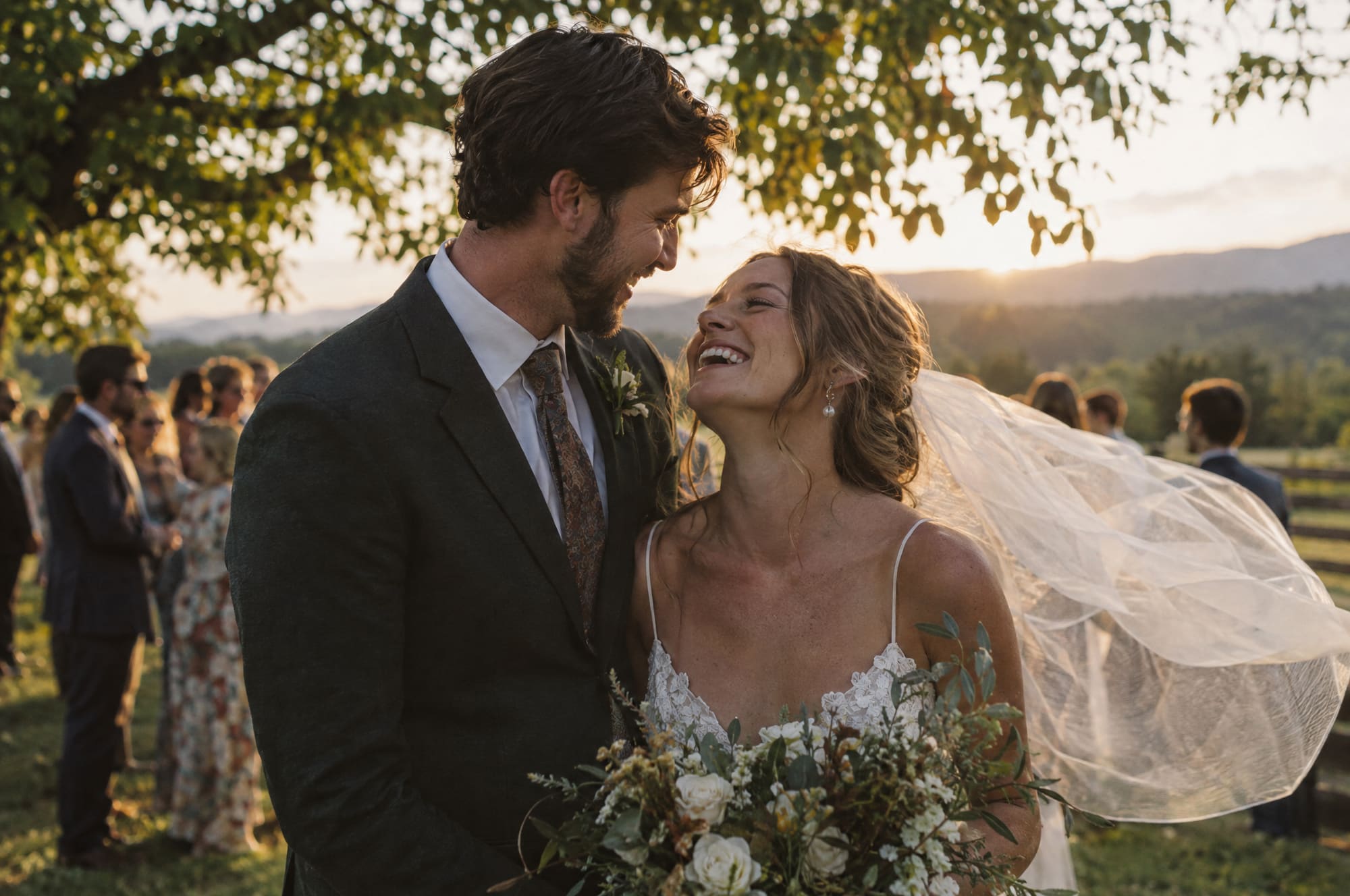 A bride laughs up at her groom under an oak at golden hour, guests and hills behind them